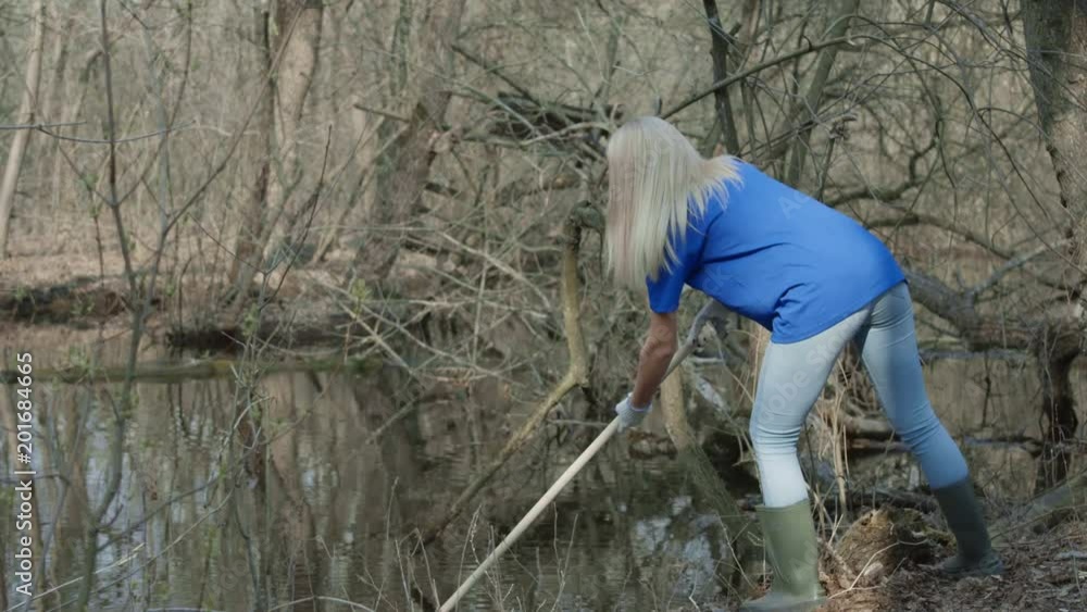 Girl in t-shirt of volunteer company cleaning up trash on pond with rake and talking to black mug managing process. Girl in t-shirt of volunteer company cleaning up trash on pond with rake and talking to black mug managing process.