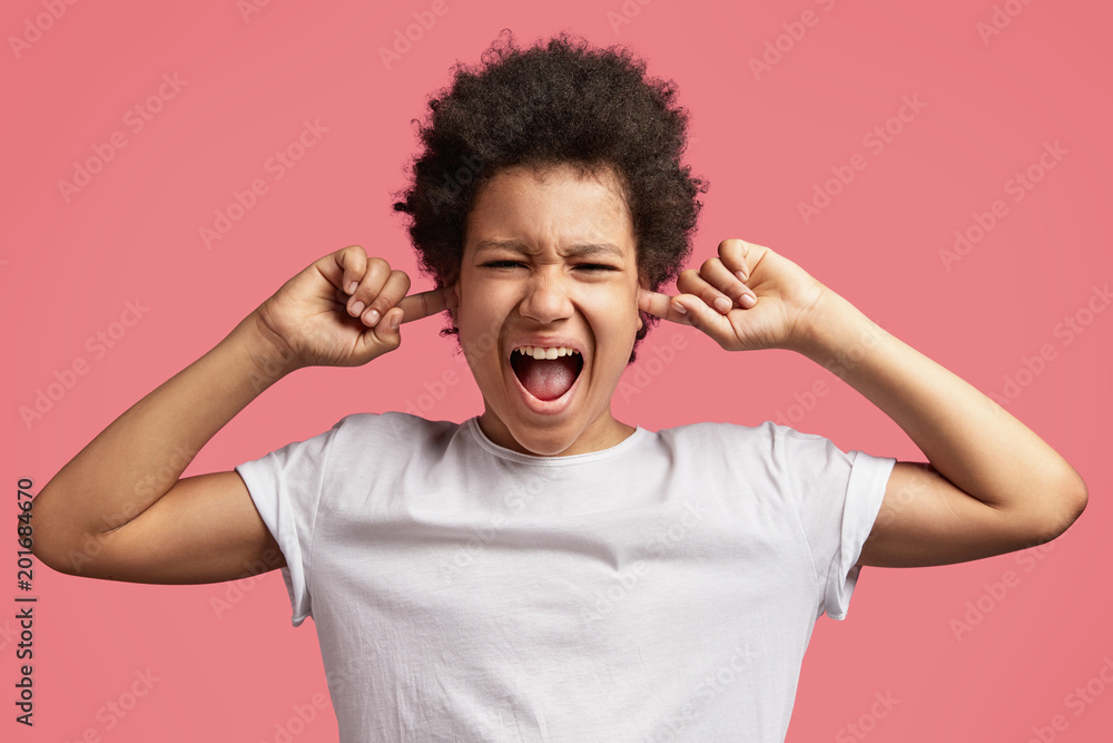 Photo of African American mad male child shouts loudly and plugs ears, hears irritated sound, has displeased expression, isolated over pink background. Mixed race teenager with discontent look Photo of African American mad male child shouts loudly and plugs ears, hears irritated sound, has displeased expression, isolated over pink background. Mixed race teenager with discontent look