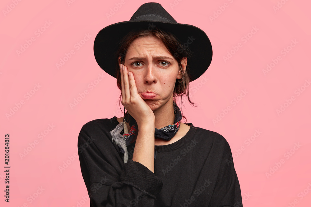 Upset beautiful female with mournful expression, curves lips and holds chin, has sullen gloomy look, wears cowboy`s stylish clothes, feels frustrated and tired after work at farm, isolated on pink Upset beautiful female with mournful expression, curves lips and holds chin, has sullen gloomy look, wears cowboy`s stylish clothes, feels frustrated and tired after work at farm, isolated on pink