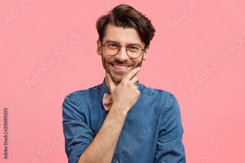 Portrait of happy bearded young male has trendy hairdo, wears fashionable jean shirt, holds chin, looks with delighted cheerful expression at camera, satisfied to communicate with beautiful woman.