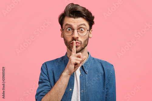 Horizontal portrait of amazed bearded male with trendy hairdo, keeps index finger on lips, stares as afraids his secret will reveal to all people, asks keep silence, isolate over pink background