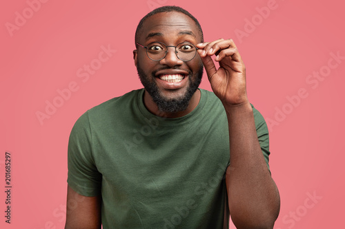Glad dark skinned man with broad smile, has white perfect teeth, looks attentively through spectacles, glad to hear interesting story, poses against pink background. Delighted Africaan American male