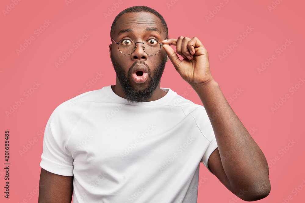 Portrait of handsome African American young male manager looks with big surprisment on workload, realizes that he has deadline, dressed in casual white t shirt, isolated over pink background. Portrait of handsome African American young male manager looks with big surprisment on workload, realizes that he has deadline, dressed in casual white t shirt, isolated over pink background.