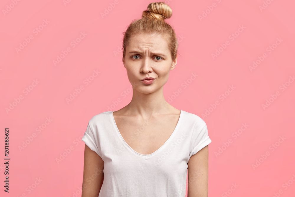 Human facial expressions concept. Attractive lovely young female model with light hair tied in knot, frowns lips in displeasure, feels puzzlement, can`t find solution in mind, isolated over pink wall Human facial expressions concept. Attractive lovely young female model with light hair tied in knot, frowns lips in displeasure, feels puzzlement, can`t find solution in mind, isolated over pink wall