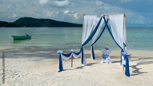 Beautiful tropical beach with decorated wedding pergola with blue and white materials prepared for ceremony. In the centre visible table with champagne. 