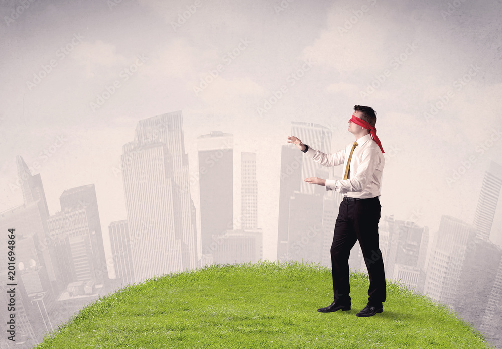 Young blindfolded businessman steps on a a patch of grass with a cloudy city in the background Young blindfolded businessman steps on a a patch of grass with a cloudy city in the background
