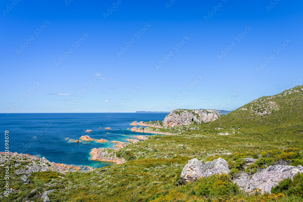 Stunning view from bright white light station to deep blue sea ocean bay turquoise water with orange red rocks at shore coast on warm sunny clear sky day, Rocky Cape National Park, Tasmania, Australia