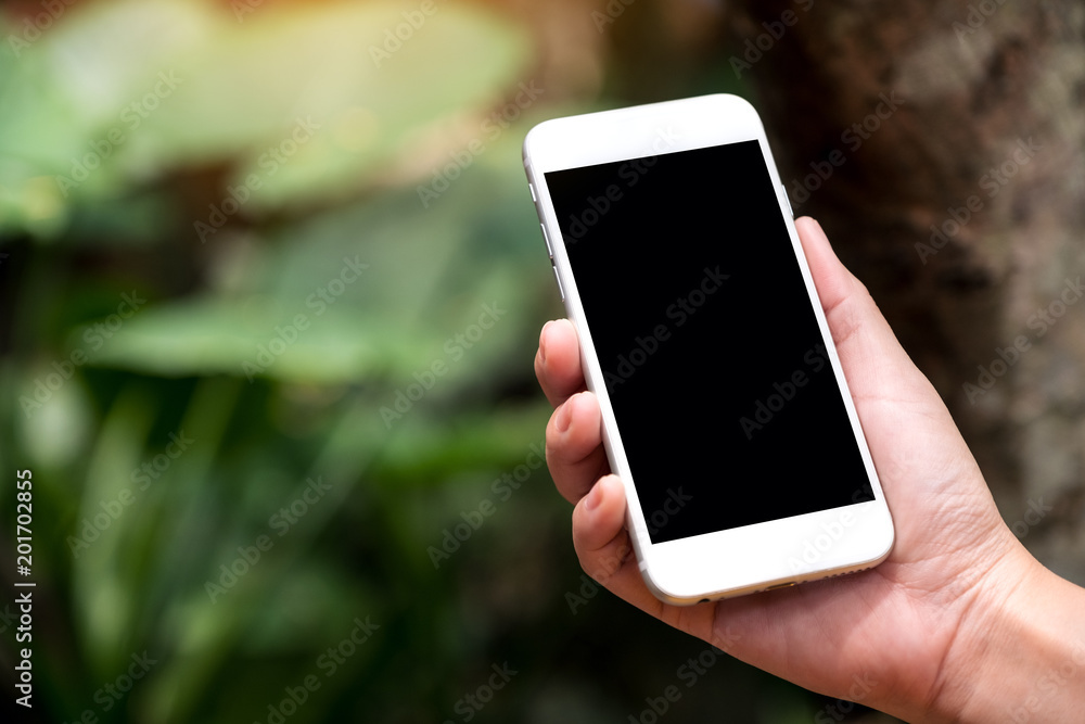 Mockup image of a hand holding white smart phone with blank black desktop screen in outdoor with blur green nature background