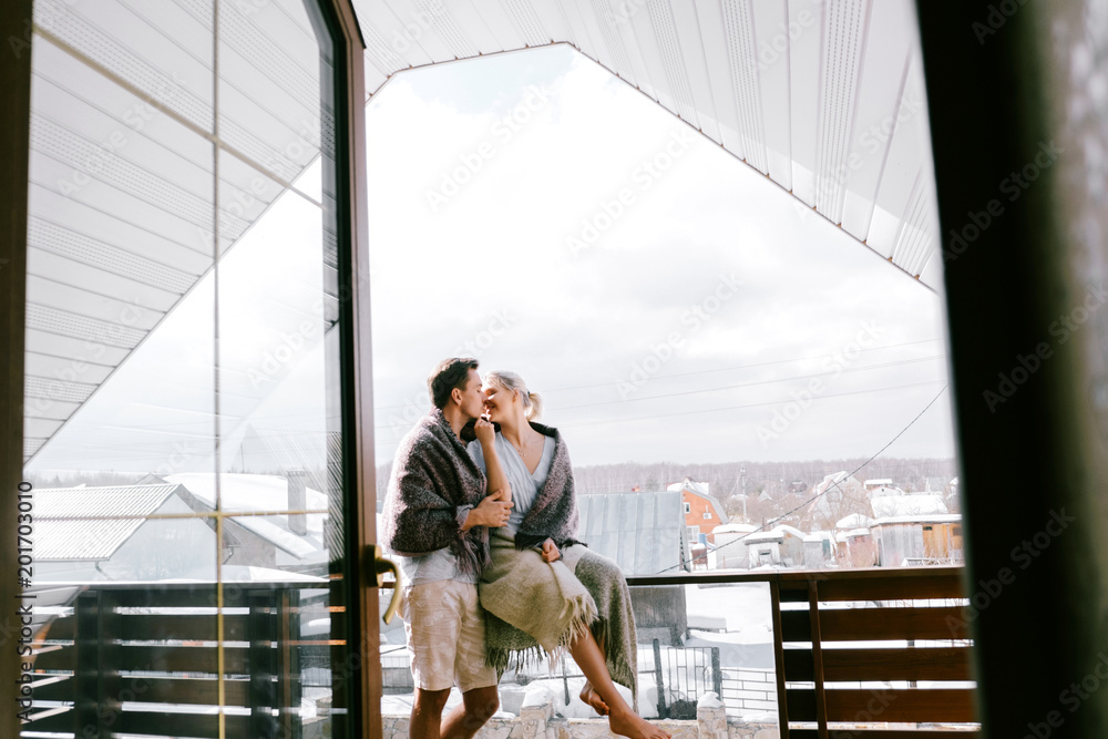 Lovely couple together at home. Pretty woman and attractive man wearing stylish casual clothes and blanket standing on a balcony and hugging Lovely couple together at home. Pretty woman and attractive man wearing stylish casual clothes and blanket standing on a balcony and hugging