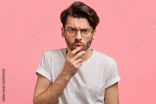 Portrait of bearded young stylish male looks with serious attentive expression at camera, tries to see something, has puzzled surprised look, wears casual t shirt, isolated over pink background.