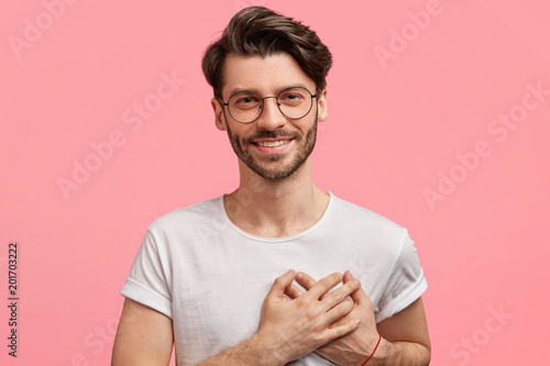 Pleased attractive male model with appealing look, has stylish haircut and bristle, keeps hands on chest, expresses his truthful feeling to girlfriend, has positive smile on face, demonstrates love © wayhome.studio 