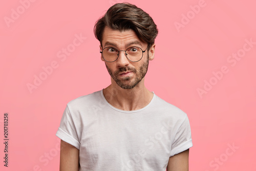 Horizontal portrait of serious unshaven male manager looks with thoughtful hesitant expression, curves lips, tries to find solution in mind, wears casual white t shirt, isolated over pink background