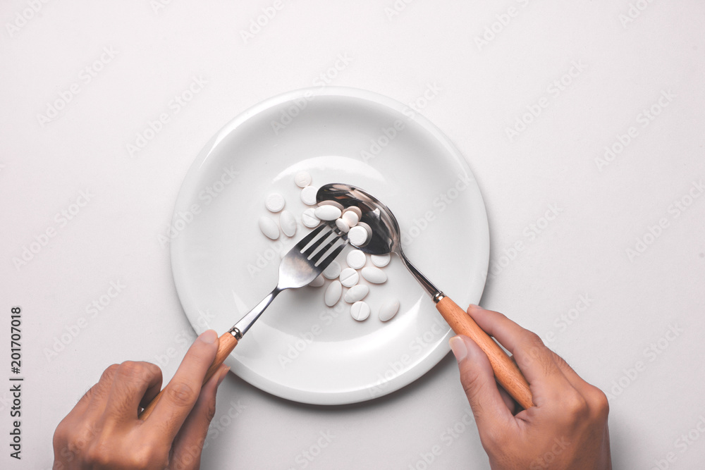 Φωτογραφία Top view of man's hands at dining table holding a fork and knife above dish with pills over pink background