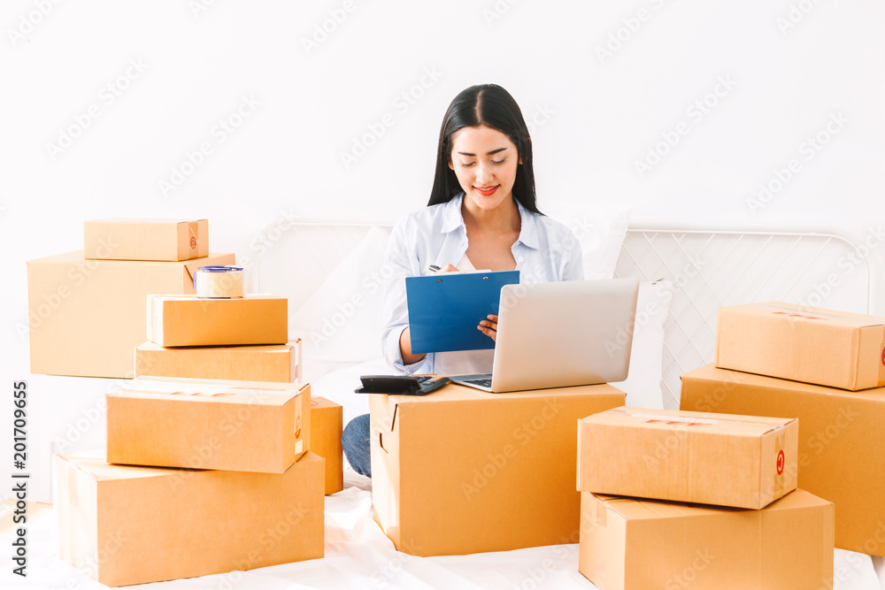 Young woman freelancer working and checklist and writing order with cardboard box on bed at home - SME business online and delivery concept Young woman freelancer working and checklist and writing order with cardboard box on bed at home - SME business online and delivery concept