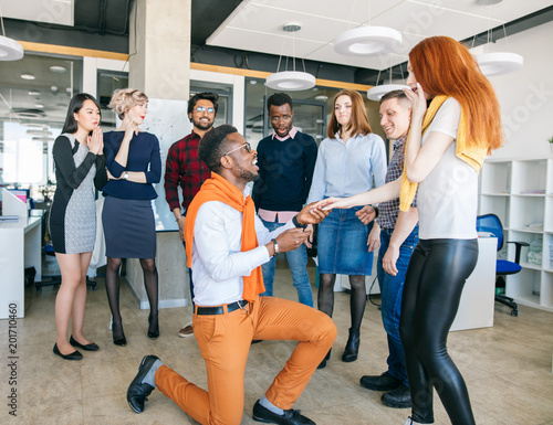 Handsome African man downing on bended knee and proposing marriage to a beautiful sirprised ginger woman in the modern office in front off employees photo