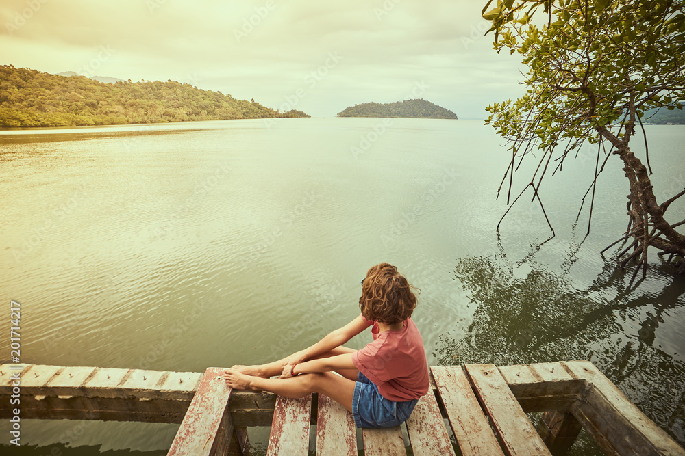 Fototapeta premium Traveling girl on the wood pier. Pretty young woman and tropical landscape. Summer lifestyle and adventure photo. Fish eye lens image