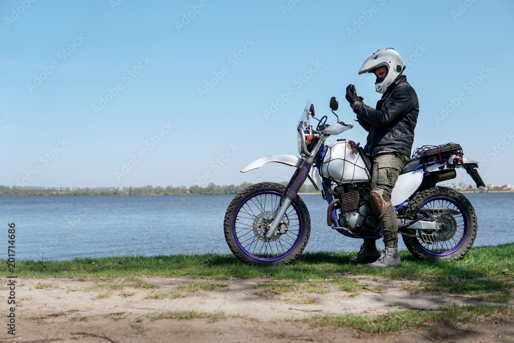 Young stylish man sit on classic retro off road track motorcycle on the beach, outdoor portrait, posing, in lather jacket and Sunglasses, travel active lifestyle concept, ocean, sea, lake, river