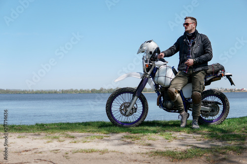 Young stylish man sit on classic retro off road track motorcycle on the beach  outdoor portrait  posing  in lather jacket and Sunglasses  travel active lifestyle concept  ocean  sea  lake  river