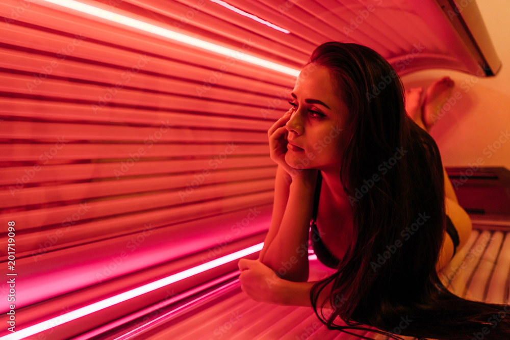 a young long-haired girl lies in a horizontal solarium, sunbathing under ultraviolet rays - obrazy, fototapety, plakaty 