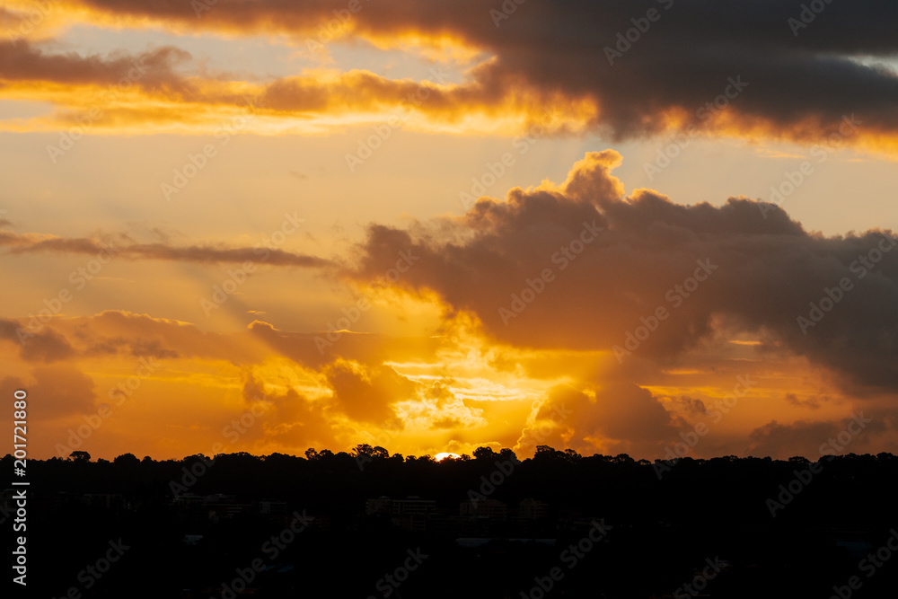 Orange glowing sunset with rays emanating through dark clouds over a silhouetted landscape. Perth, Western Australia.