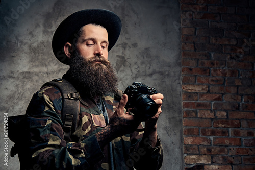 Portrait of a handsome bearded traveler in a hat and camouflage jacket, with a backpack and tattooed arms, holds a photo camera, isolated on a dark background.