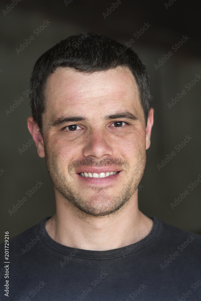 Portrait of young handsome black-haired unshaven confident successful man in casual T-shirt with white teeth and shiny black eyes smiling happily in camera on dark background. Portrait of young handsome black-haired unshaven confident successful man in casual T-shirt with white teeth and shiny black eyes smiling happily in camera on dark background.