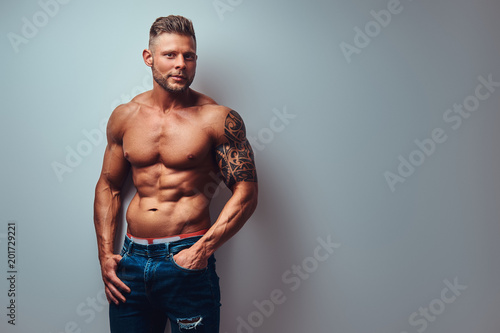 A handsome shirtless tattooed bodybuilder with stylish haircut and beard, wearing sports shorts, posing in a studio. Isolated on a gray background