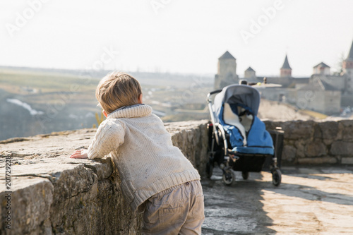 Back view of the little boy who is standing against the background of ancient castle outdoor and looking at it
