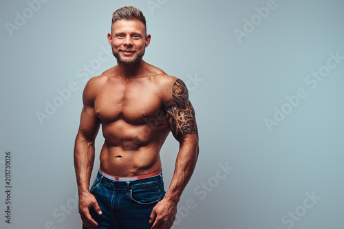 A handsome shirtless tattooed bodybuilder with stylish haircut and beard, wearing sports shorts, posing in a studio. Isolated on a gray background