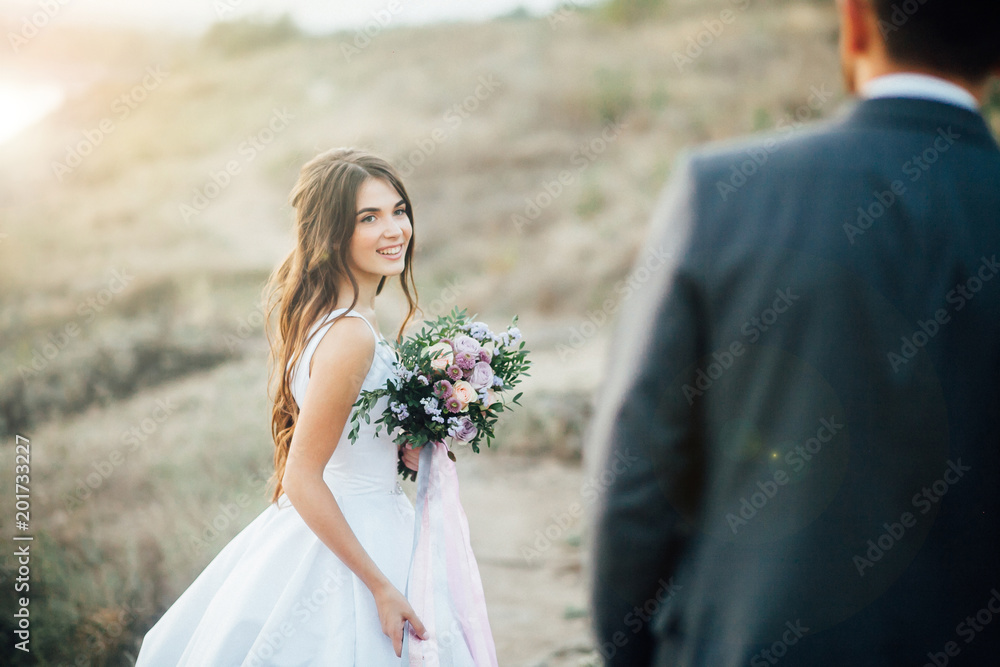 Young bride with wedding bouquet at the hands looking to groom with smile at sunset. Outdoors Young bride with wedding bouquet at the hands looking to groom with smile at sunset. Outdoors