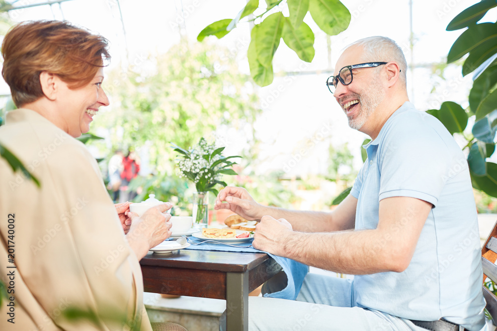 Laughing senior wife and husband sitting by served table by breakfast and having talk Laughing senior wife and husband sitting by served table by breakfast and having talk