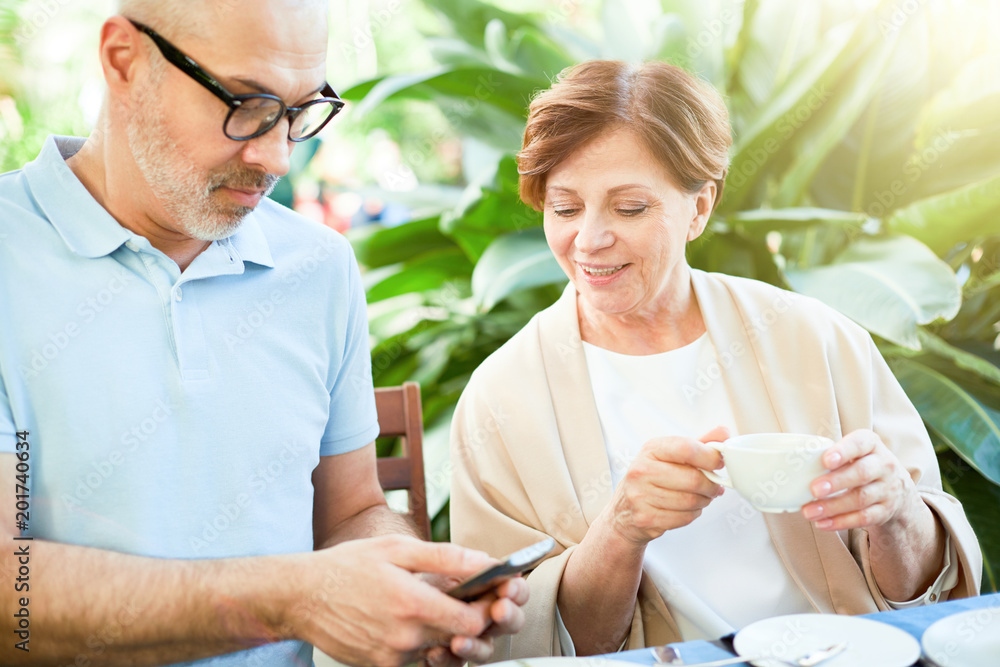 Aged man with smartphone showing message or notification to his wife by breakfast in orangery Aged man with smartphone showing message or notification to his wife by breakfast in orangery
