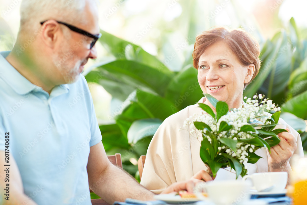 Mature woman with bunch of fresh lilies-of-the-valley talking to her husband by breakfast in home garden