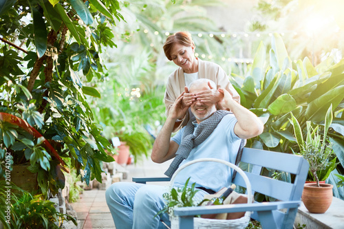Photos Playful senior woman covering eyes of her husband by hands while both having res