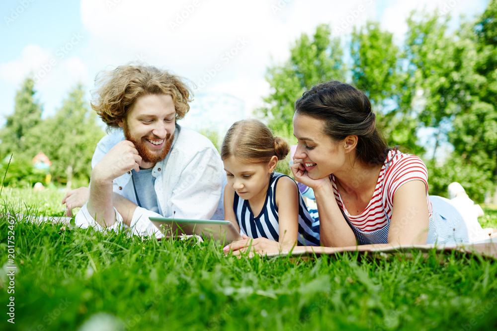 Fototapeta premium Contemporary family of father, mother and daughter with tablet lying on green lawn, relaxing and watching video in the net