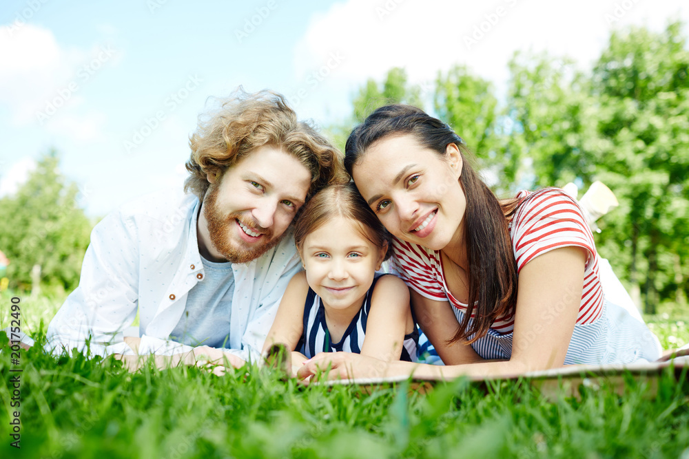 Fototapeta premium Young couple and their daughter looking at camera while relaxing on green lawn in natural environment
