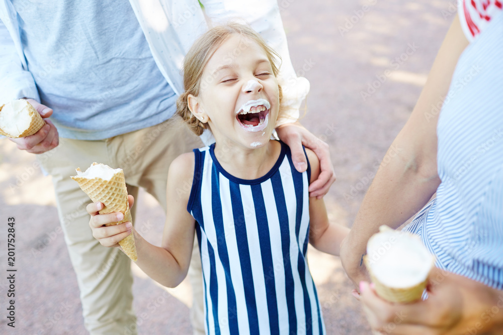 Laughing girl with grimy face eating ice cream and enjoying promenade with her parents on sunny day