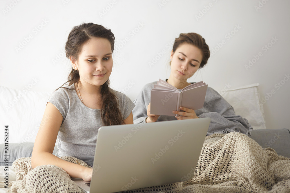 Positive teenage girl dressed casually doing homework using high speed internet connection on generic laptop computer in bedroom while her elderly sister sitting behind her, absorbed in book
