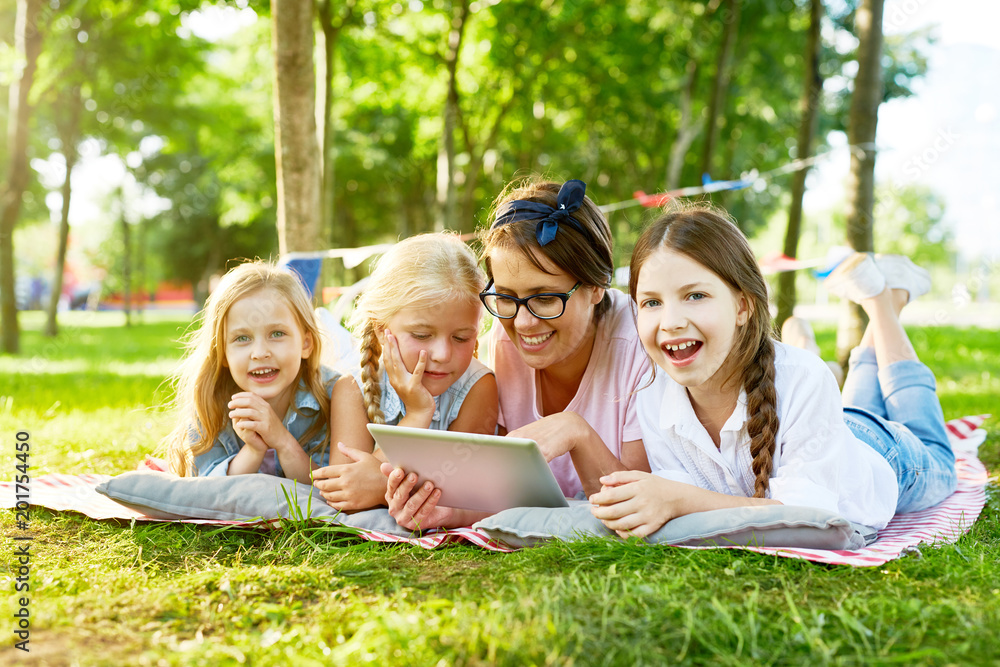 Fototapeta premium Happy little girls and their teacher with tablet spending weekend in park by watching movie or videos in the net