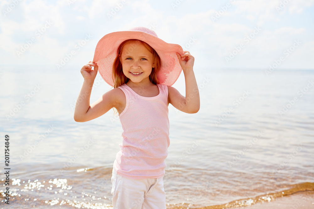 Little girls in beautiful hat standing by waterside and posing for camera on hot summer day during vacation Little girls in beautiful hat standing by waterside and posing for camera on hot summer day during vacation