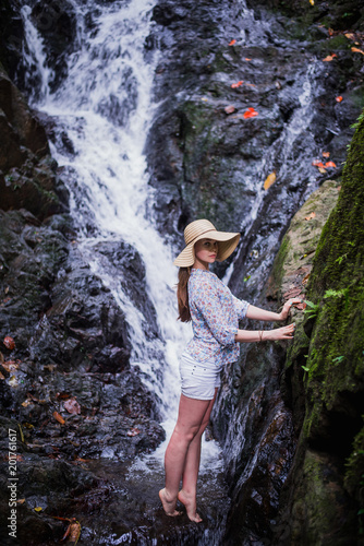 Tropical traveling. Young woman in hat enjoying waterfall view.