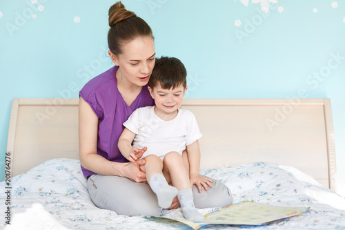 Indoor shot of affectionate young mother holds and embraces her small son, look attentively at book with colourful pictures, read interesting stories for children, sit on soft comfortable bed photo