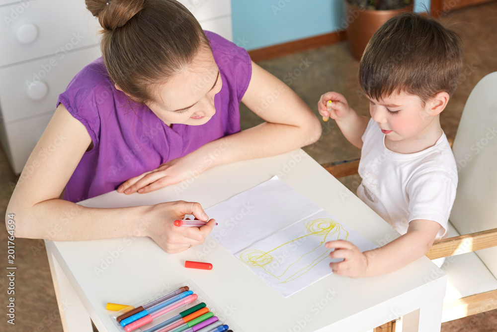 Top view of clever talented small boy draws car on paper with colourful mark pen, likes to make different pictures, spends time with his affectionate mother. Children and development concept Top view of clever talented small boy draws car on paper with colourful mark pen, likes to make different pictures, spends time with his affectionate mother. Children and development concept