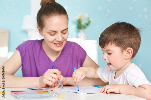 Photo of talented diligent small boy draws with great desire picture, uses colourful marker, cheerful mother helps to choose colors, sit together against blue wall in room. Creativity and art
