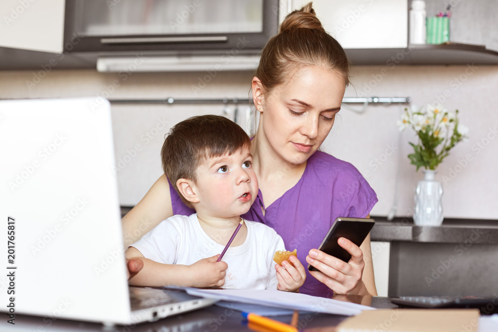 Photo of busy working mother tries to solve financial problems, waits for call, holds smart phone, sits with child at work place, earns money on maternity leave, surrounded with papers. Distance work Photo of busy working mother tries to solve financial problems, waits for call, holds smart phone, sits with child at work place, earns money on maternity leave, surrounded with papers. Distance work