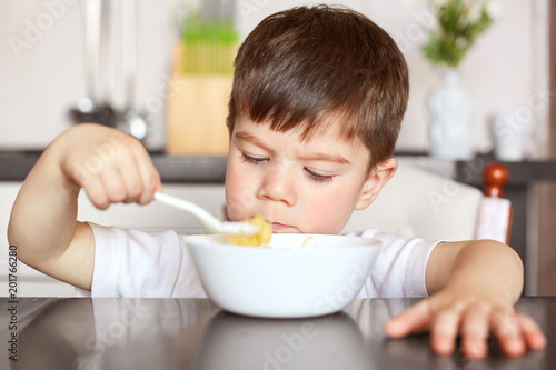Healthy eating and children concept. Handsome small child eats with great appetite delicious porridge prepared by mother, holds big spoon and looks at bowl, sits against domestic kitchen interior