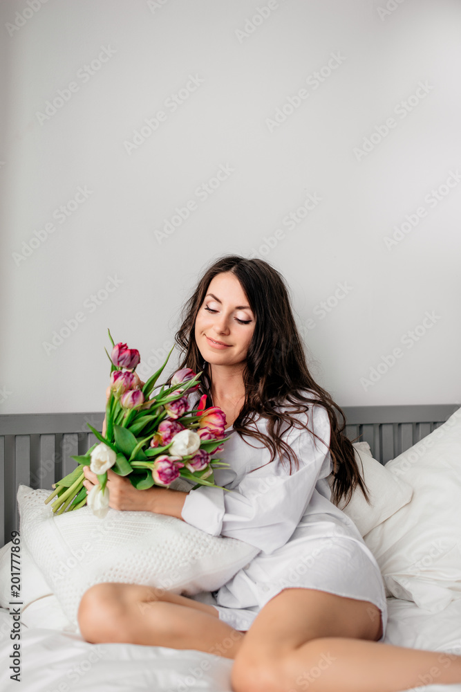 beautiful young brunette girl in white shirt with bouquet of tulips on bed at home beautiful young brunette girl in white shirt with bouquet of tulips on bed at home