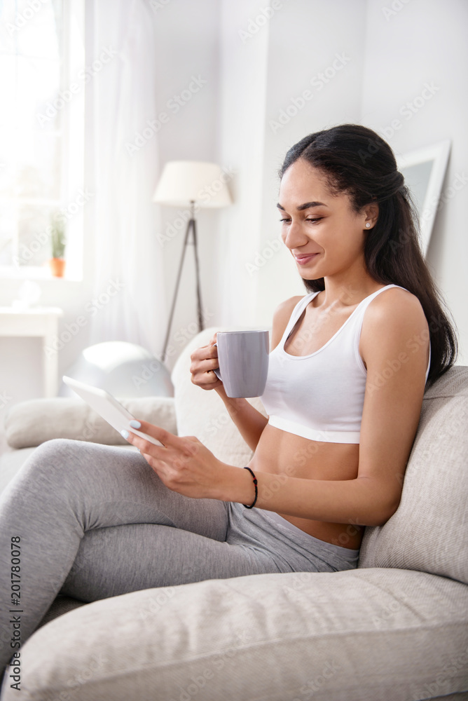Enjoying free time. Pleasant athletic woman sitting on the sofa and reading from tablet and smiling cutely while drinking tea Enjoying free time. Pleasant athletic woman sitting on the sofa and reading from tablet and smiling cutely while drinking tea