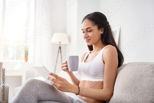 Cozy reading. Pleasant young woman sitting on the sofa and reading from tablet while drinking tea and smiling
