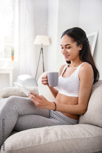 Enjoying free time. Pleasant athletic woman sitting on the sofa and reading from tablet and smiling cutely while drinking tea